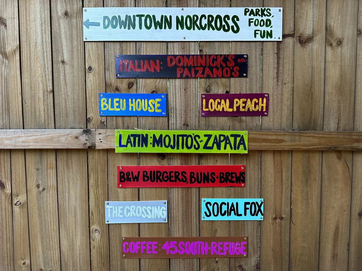 Hand-painted wooden signs on the fence pointing to downtown Norcross, naming nearby restaurants and a coffee shop