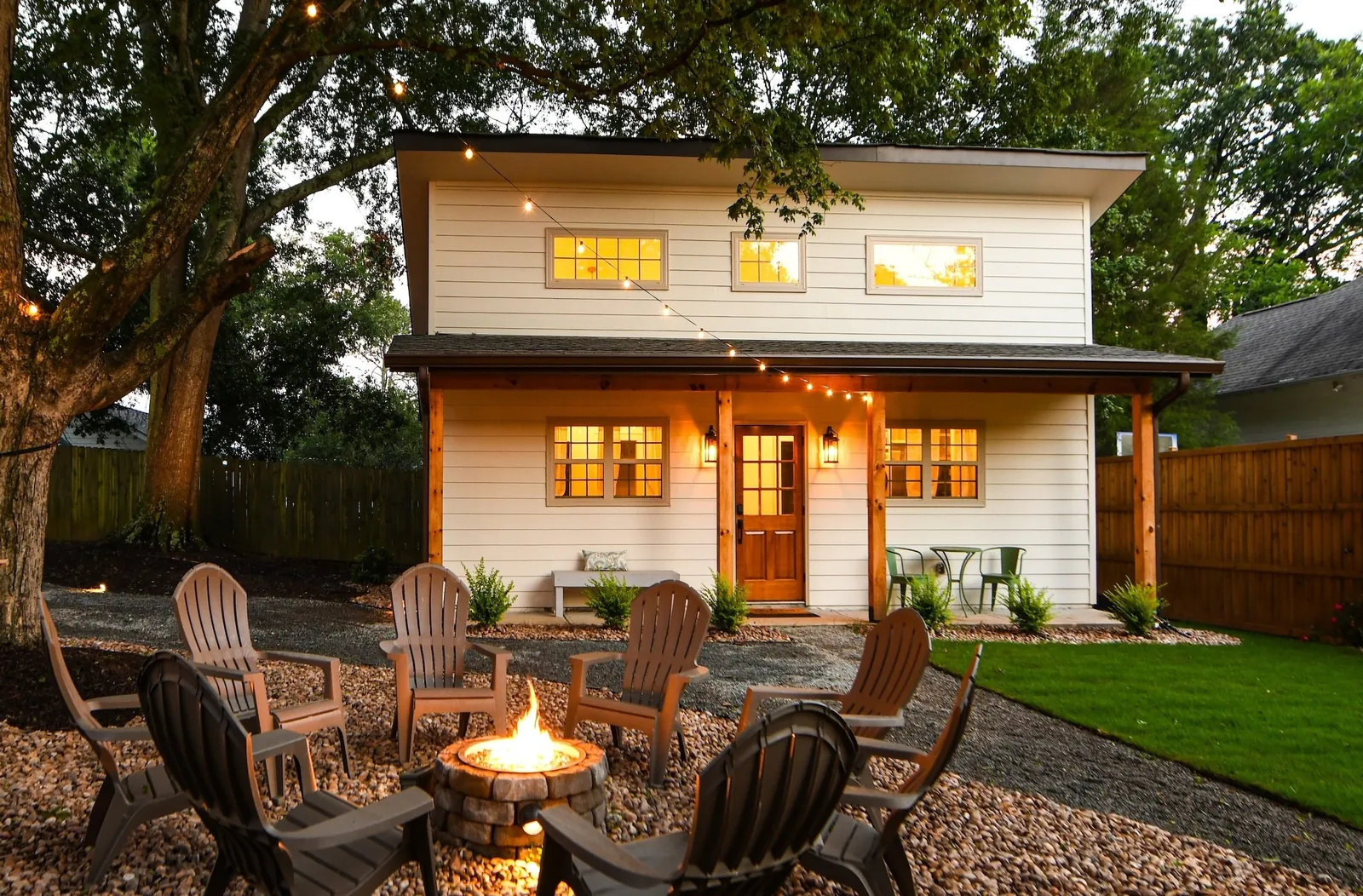 The two-story Cozy Cottage at dusk behind the lit gas fire pit and Adirondack chairs, with string lights and a hammock in the shared yard